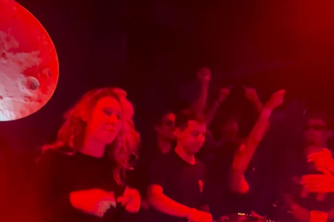Duas pessoas atrás dos CDJs, com a lua do Moon Club atrás e o público de braços no ar sob luz vermelha