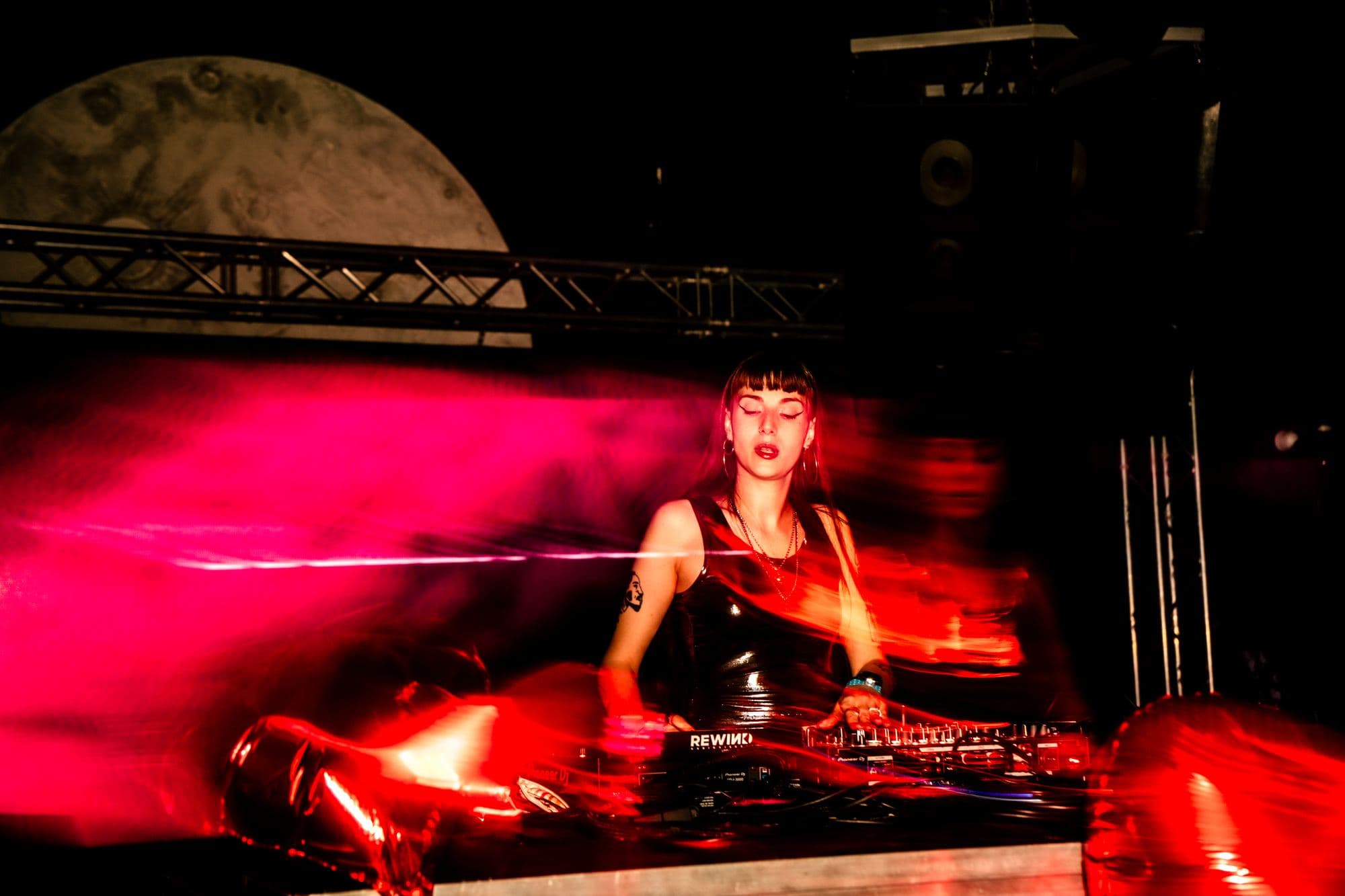Palco do Moon Club com a lua gigante atrás de uma DJ, em luz vermelha